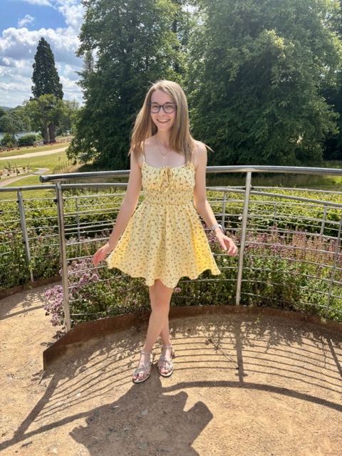 A young woman in a yellow floral dress smiles while standing on a circular metal-railed platform surrounded by green plants and flowers on a sunny day.