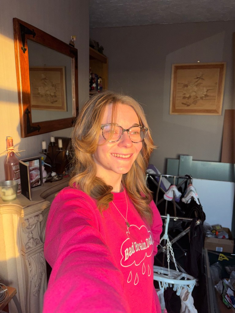 A person smiling while taking a selfie, wearing a pink sweatshirt with the text 'Bad Brain Dad.' The room in the background features wooden frames, a shelf, and some laundry drying equipment.