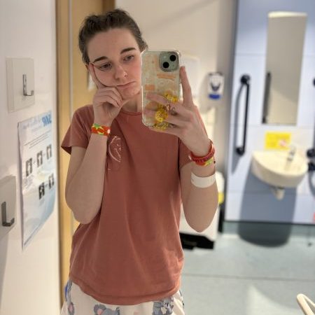 A person wearing a pink t-shirt and pajama pants with koala patterns, posing for a selfie in a hospital setting with a thoughtful expression.