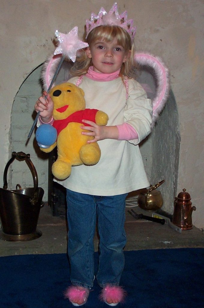 A young girl wearing a pink crown and fairy wings, holding a wand and a Winnie the Pooh stuffed toy, standing on a blue carpet near a fireplace.